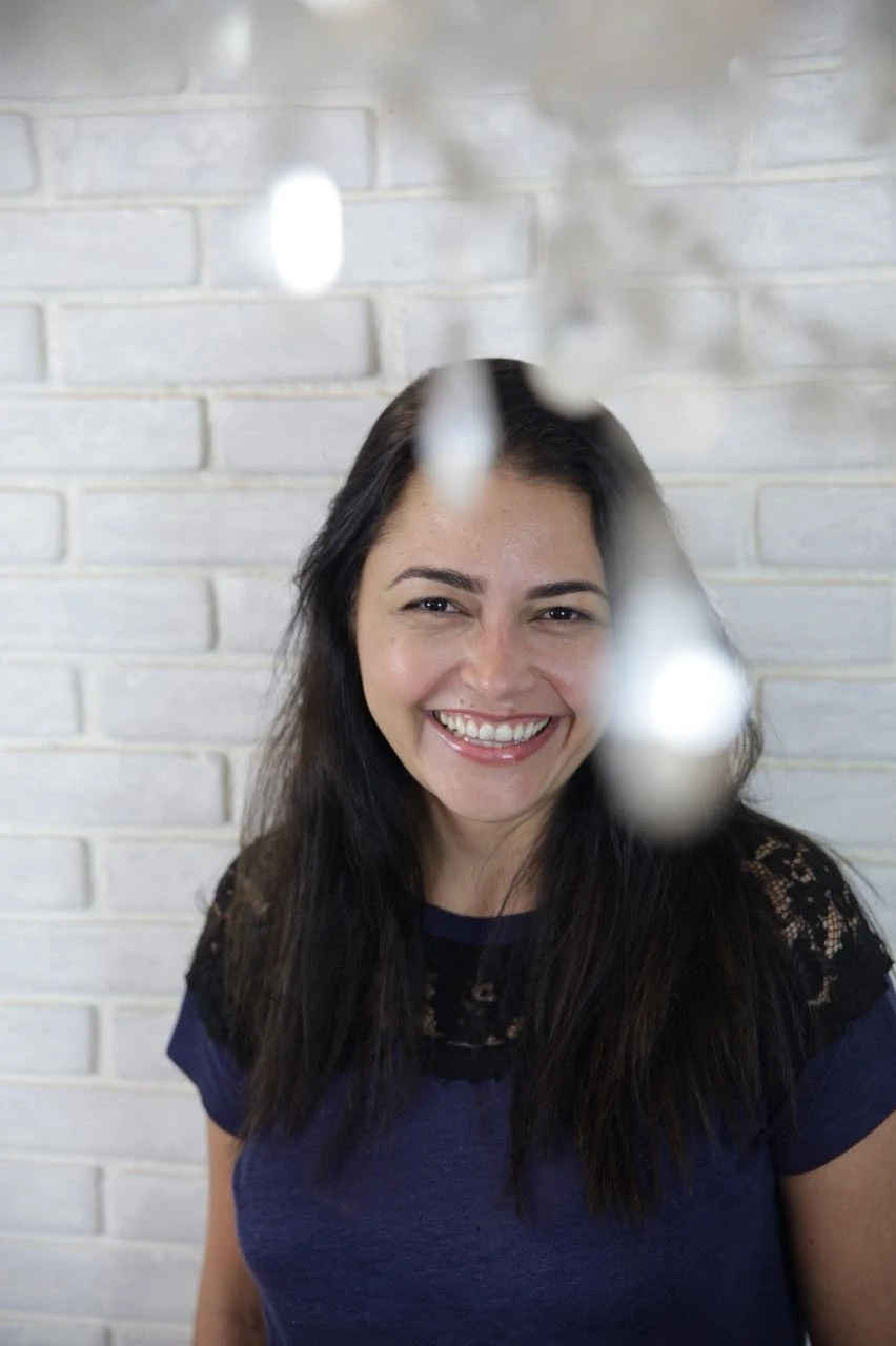Retrato de Juliana sorrindo diante de uma parede de tijolos brancos.
